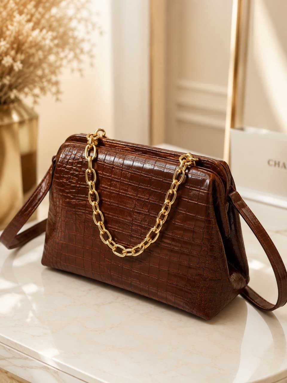 Bolsa em couro legítimo com textura croco e corrente dourada, sobre bancada de mármore